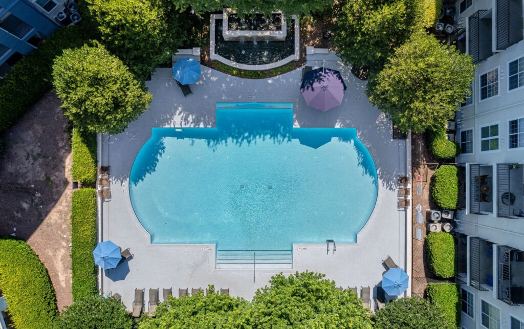 Aerial view of a large resort-style swimming pool surrounded by lounge chairs and colorful umbrellas. The pool area is framed by manicured greenery and apartment buildings, with a decorative water feature and fountain visible at the top. The bright blue water, geometric pool shape, and symmetrical layout create an inviting and luxurious atmosphere.