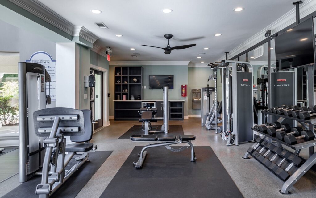Fully equipped apartment fitness center with strength training machines, free weights, benches, and cable stations. The space features a mounted TV, ceiling fan, and built-in shelving with decorative accents. Neutral-colored walls, ample lighting, and mirrored walls create a clean, spacious, and functional workout environment.