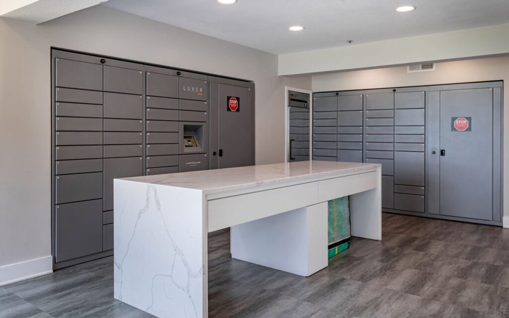 Modern package locker room featuring sleek gray Luxer One lockers with a touchscreen interface for secure parcel retrieval. A white marble-topped central table provides a surface for sorting or organizing packages. The space has neutral walls, recessed lighting, and wood-look flooring, creating a clean and organized atmosphere.