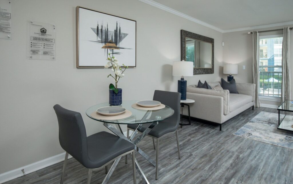 Apartments Near Perimeter Center District - The Stratford - Open Living And Dining Area With Round Glass Table, Two Modern Chairs, Sofa Seating, Large Window Light, And Wood-Inspired Flooring