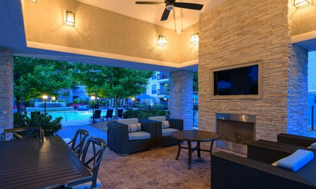 Covered outdoor lounge area at an apartment community featuring cushioned wicker furniture, a mounted flat-screen TV above a stone fireplace, ceiling fans, and ambient lighting. Beyond the patio, a swimming pool with lounge chairs is visible, surrounded by trees and landscaped greenery.
